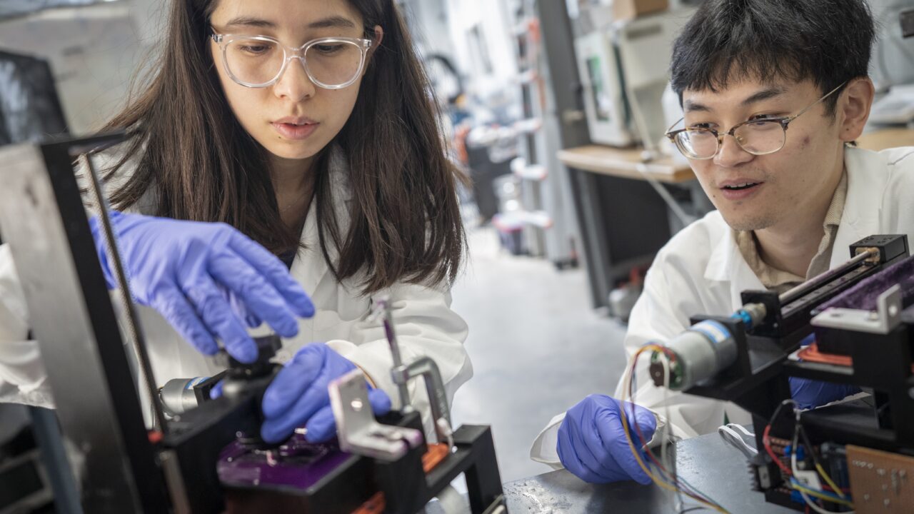 [Photo] Two students wearing lab coats and gloves adjust research equipment together in a laboratory. Instruments, cables and scientific devices surround the workstation as they focus on the experiment.
